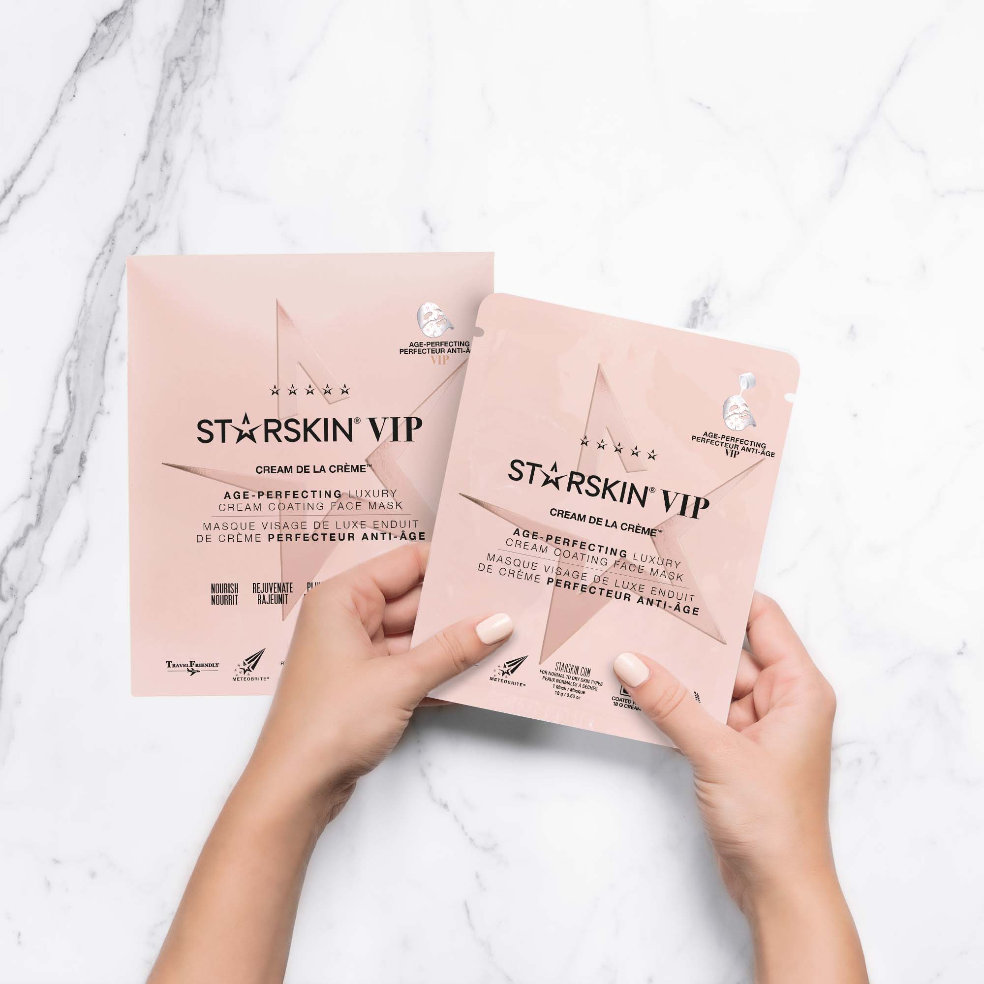 The starskin cream de la creme mask sachet is being held by someone. Underneath the sachet is the normal product packaging. The image has a marble background. 