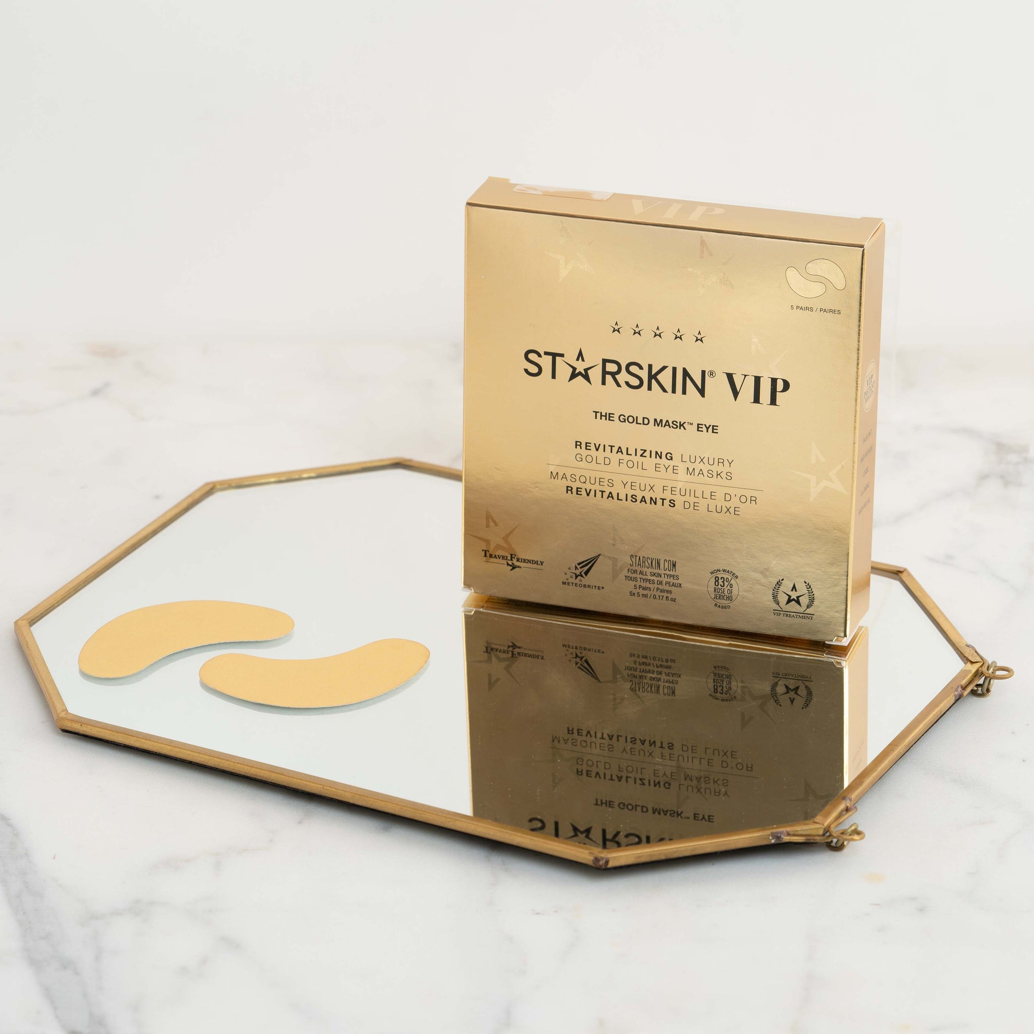 Starskin gold eye mask product packaging on top of a small mirror. Next to the packaging on the mirror are the two eye patches. The mirror is laying on a marble background.