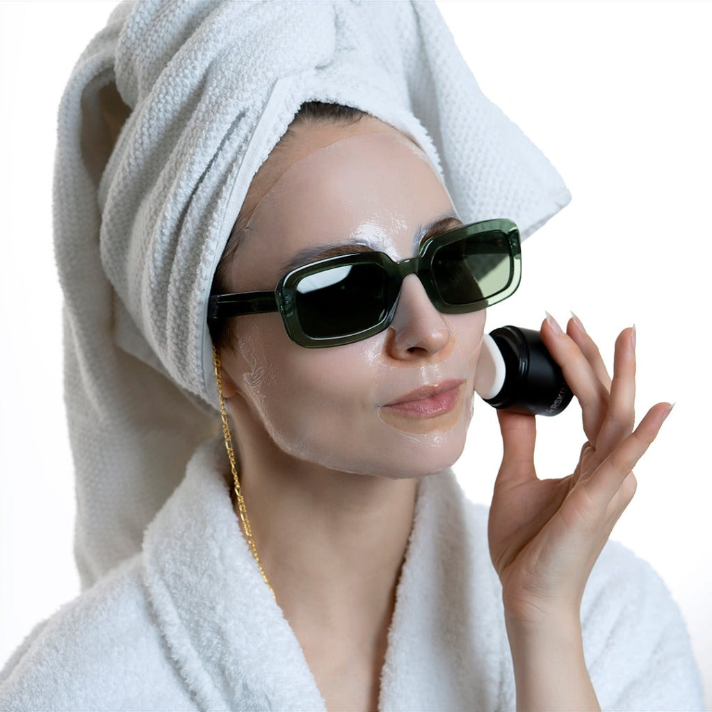 Image of a model wearing a bathrobe, a towel on her hair and green sunglasses. She is wearing a STARSKIN Coconut Bio-Cellulose mask on her face and she is holding the STARSKIN Artist FX with the Artist FX Ceramic Stone against the mask