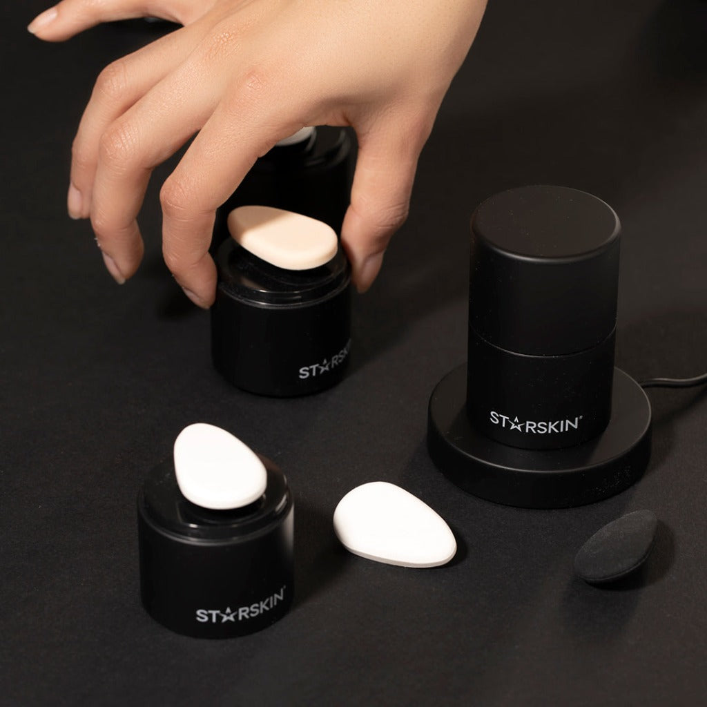 Image with black background showing a STARSKIN Artist FX standing on the charger. Next to it are two more STARSKIN Artist FX with a Ceramic stone and a Silicone puff. Around it is a Rubycell puff and a Ceramic Stone. A hand entering from above the image grabs a STARSKIN Artist FX with the Silicone puff